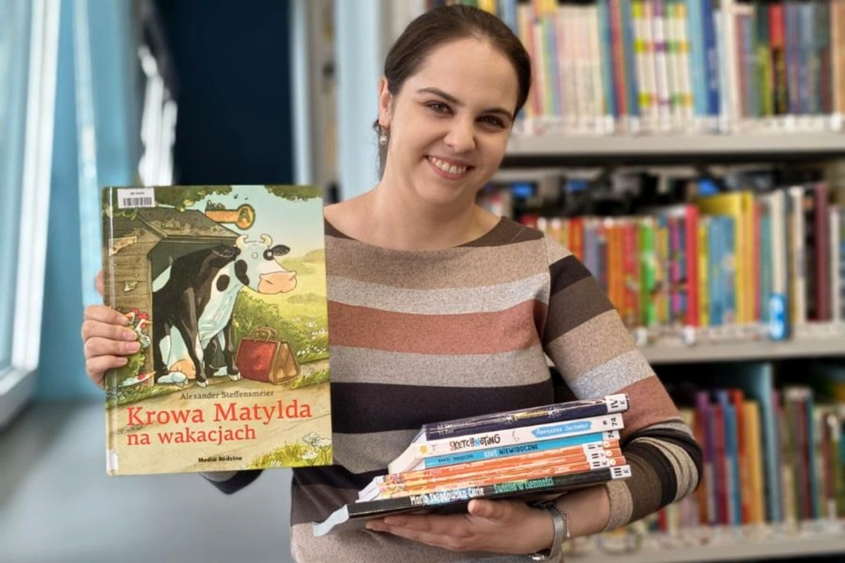 Uśmiechnięta kobieta w swetrze w paski trzyma książkę „Krowa Matylda na wakacjach” oraz stos kolorowych książek dla dzieci. Za nią regały pełne książek.
